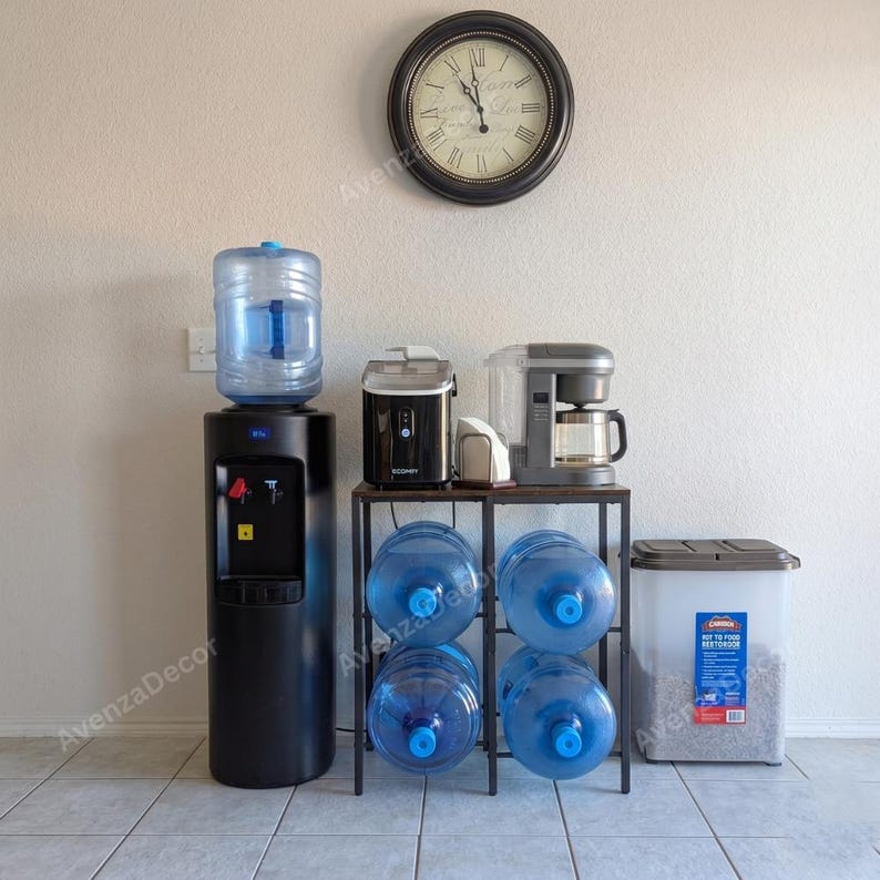 May include: A black water cooler with a large blue jug, a metal rack holding four additional blue water jugs, a black ice maker, a grey coffee maker, and a large rectangular plastic storage container with a lid, all beneath a round wall clock.