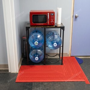May include: A red Galanz microwave sits on a black metal stand with three large clear plastic water jugs stored on the shelves. A roll of white paper towels stands next to the microwave. A red rectangular rubber floor mat lies in front.