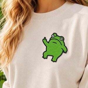May include: A cream-colored sweatshirt with a green frog patch. The frog is embroidered with a black outline and is giving the middle finger. The frog patch is on the left side of the chest.