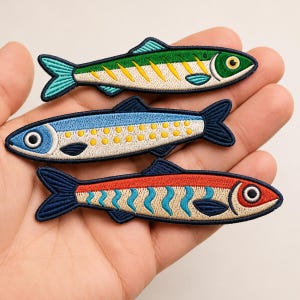 May include: Three embroidered fish patches featuring detailed stitching. The top fish is green with yellow stripes, the middle fish is blue with yellow dots, and the bottom fish is red with blue wavy lines. Each fish has a dark blue outline.