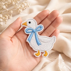May include: Embroidered white goose patch with a light blue bow. The goose has yellow feet and a yellow beak. The patch is designed with a stitched outline and is likely for application on clothing or accessories.