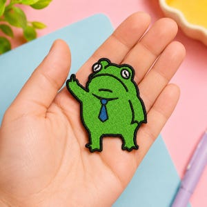 May include: Embroidered green frog patch with a blue tie, giving a hand gesture. The frog has white eyes and a black outline. The patch is held in a hand against a light blue and pink background.