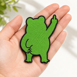 May include: A green embroidered patch featuring a cartoon frog with a black outline. The frog is giving the middle finger with one hand and has its other hand on its rear. The patch is held in a person's hand.