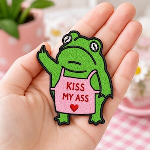 May include: Embroidered patch featuring a green frog with a pink apron. The apron has the text "KISS MY ASS" and a red heart. The frog is giving the middle finger.