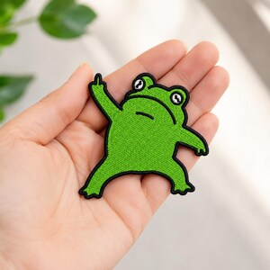May include: Embroidered green frog patch with black outline, raising a single finger. The frog has large, round eyes and a grumpy expression. This patch is a fun accessory for jackets, bags, or other fabric items.