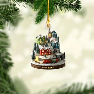 May include: A hanging Christmas ornament featuring a miniature winter village scene with colorful houses, snow-covered pine trees, and a small train on a track. The base of the ornament includes the text YOUR NAME in gold lettering on a dark brown rim.
