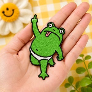May include: Embroidered green frog patch with a white undergarment, extending a middle finger. The frog has a pink tongue sticking out and is wearing a white undergarment. The patch is held in a hand.