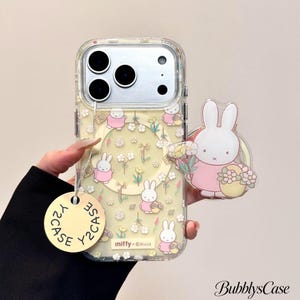 Op de afbeelding: Een doorzichtig smartphonehoesje met een herhalend patroon van witte Miffy-konijntjes in roze jurkjes en kleine gele bloemetjes. Het hoesje bevat een cirkelvormige magnetische ring, een Miffy-telefoonhouder en een rond beige label Y2CASE.