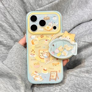 Może przedstawiać: Jasnoniebieskie i żółte etui na telefon z motywem kotów i chleba. Etui zawiera ilustracje chleba, pączków i kotów. Do etui przymocowany jest wyskakujący uchwyt w kształcie ryby z kotem i jedzeniem.