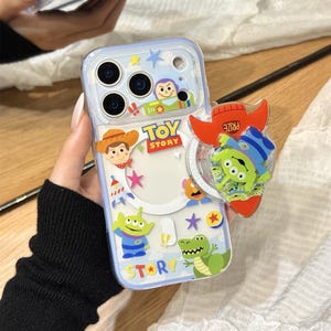 Puede incluir: Funda transparente para teléfono con personajes de Toy Story, incluyendo a Woody, Buzz Lightyear y los pequeños hombrecillos verdes. La funda tiene un borde azul y un pop socket en forma de cohete. El texto "TOY STORY" y "STAR" son visibles.