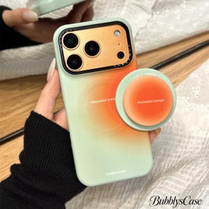 May include: A light green smartphone case with a central orange gradient circle, featuring the text "Minimalist orange" and "CNISS KAHEY". It includes a matching circular phone grip with the text "LOVELY POWER" and "Minimalist orange".