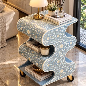 May include: A light blue, S-shaped side table featuring an intricate white floral and vine inlay pattern. The table has three tiers for storage, gold-toned metal casters for mobility, and a smooth, glossy finish across its curved, wave-like structure.