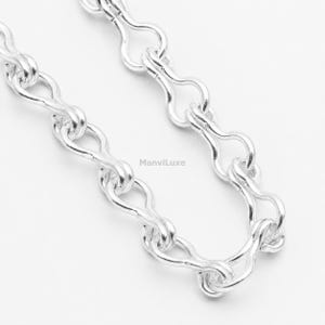 May include: A silver-toned chain necklace featuring a series of elongated, rounded links that create a continuous, flowing pattern. The metal surface is polished and reflective. The text ManviLuxe is printed in small, dark letters on the chain.