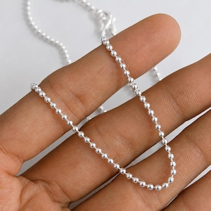 May include: A silver-toned ball chain necklace featuring a series of small, uniform spherical beads connected by thin metal links. The chain has a reflective, metallic finish and is draped in a curved line across the fingers, showing the texture.