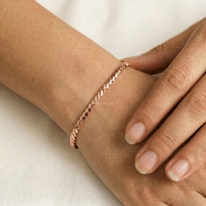 Op de afbeelding: Een roségoudkleurige kettingarmband met een reeks in elkaar grijpende, afgeplatte en gepolijste schakels. De armband wordt om de pols gedragen en toont een doorlopend, uniform ontwerp met een metallic afwerking die het licht reflecteert.