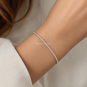 May include: A thin, silver-toned chain bracelet with a delicate, textured link design. The bracelet features a subtle, shimmering metallic finish and is worn around the wrist. The word Manviluxe is faintly visible in the center of the image.