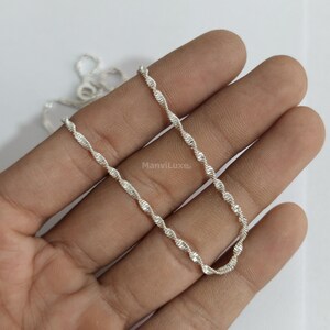 May include: A silver-colored rope chain necklace featuring a twisted, spiraling link design with a polished, reflective finish. The chain is draped in a gentle curve, showcasing the uniform, repeating pattern of the metallic links throughout the length.