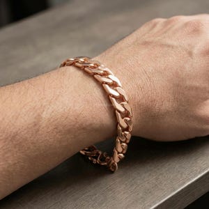 Pode incluir: Uma pulseira de corrente tipo grumet em tom de ouro rosa com elos ovais planos, polidos e entrelaçados. A pulseira tem um acabamento metálico reflexivo e é fixada com um mecanismo de fecho de gancho visível na parte inferior do pulso.