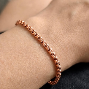 May include: A copper-coloured chain bracelet featuring a series of interconnected, rounded links. The bracelet has a metallic finish and a uniform, repeating pattern throughout its length, creating a flexible and continuous circular design.