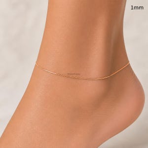 May include: A delicate rose gold anklet chain with a 1mm thickness. The fine metal chain features a simple link design that rests around the ankle. The text 1mm is visible in the upper right corner, indicating the width of the jewellery piece.