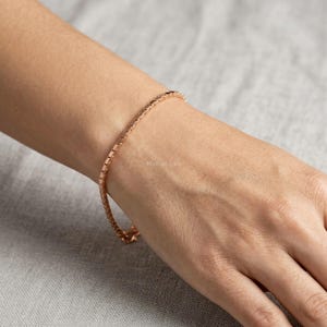 May include: A delicate rose gold chain bracelet featuring a series of small, uniform, rectangular links. The bracelet has a polished metallic finish and is secured with a small clasp, resting against the skin of a wrist with a subtle sheen.