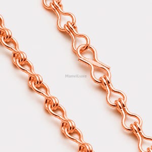Peut inclure: Une chaîne de collier en métal couleur or rose composée d'une série de maillons entrelacés en forme de huit. La chaîne présente une surface polie et réfléchissante ainsi qu'un fermoir à crochet sécurisé. Le texte ManviLuxe est visible.