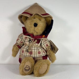 May include: A tan plush teddy bear wearing a pointed hat with a maroon band, a maroon collar, and a tan plaid shirt featuring small red leaf patterns. The bear has a black nose, a black bird accessory on its left side, and a basket icon on its foot.