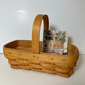 May include: A rectangular woven basket with a tall, arched handle. The basket is made of light brown wood and features a woven design. A small card with the "Longaberger" logo is inside the basket.