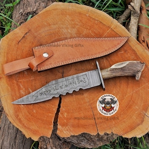 May include: A fixed-blade knife with a patterned steel blade featuring serrated edges and etched runic symbols. The handle is crafted from textured brown antler. A tan leather sheath with a metal snap button closure accompanies the knife.
