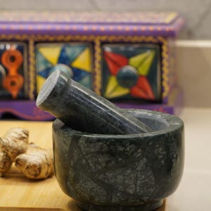 Könnte beinhalten: Ein dunkelgrüner Marmor-Mörser und Stößel-Set. Der Mörser ist rund mit breitem Boden, der Stößel liegt darin. Das Set steht auf einem Holzbrett, Ingwer ist daneben zu sehen. Im Hintergrund ein dekorativer Schrank.