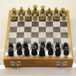 Könnte beinhalten: Ein komplettes Schachspiel mit einer Holzbox. Das Schachbrett besteht aus Marmor mit einem braun-weißen Schachmuster. Die Schachfiguren sind schwarz und grün. Das Set ist spielbereit.