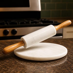 May include: A white marble rolling pin with light brown wooden handles resting diagonally across a circular white marble pastry board. The rolling pin features a smooth cylindrical barrel and tapered wooden grips on each end.