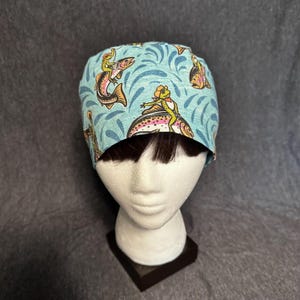 May include: A light blue surgical scrub cap featuring a whimsical design of cartoon frogs riding fish. The cap has a repeating pattern of fish and water swirls. The frogs are wearing hats and vests.
