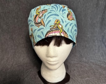 May include: A light blue surgical scrub cap featuring a whimsical design of cartoon frogs riding fish. The cap has a repeating pattern of fish and water swirls. The frogs are wearing hats and vests.