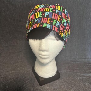 May include: A black scrub cap with the word "PRIDE" in a repeating pattern. The word is in rainbow colors with a heart symbol. The cap is displayed on a white mannequin head.