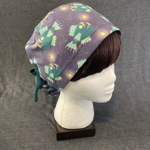 May include: A dark purple scrub hat with a green and white anglerfish print. The hat has a tie at the back.