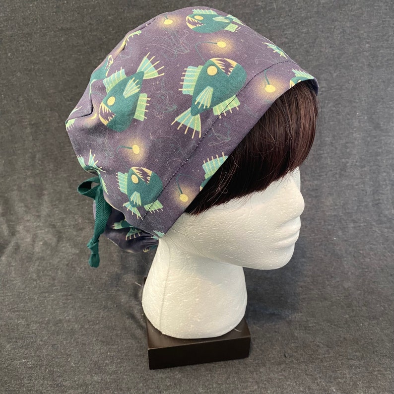 May include: A dark purple scrub hat with a green and white anglerfish print. The hat has a tie at the back.