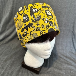 May include: A yellow and black patterned scrub cap with a repeating design of cartoon monsters, bats, and pumpkins. The cap is made of fabric and is designed to be worn on the head. The cap is shown on a mannequin head.