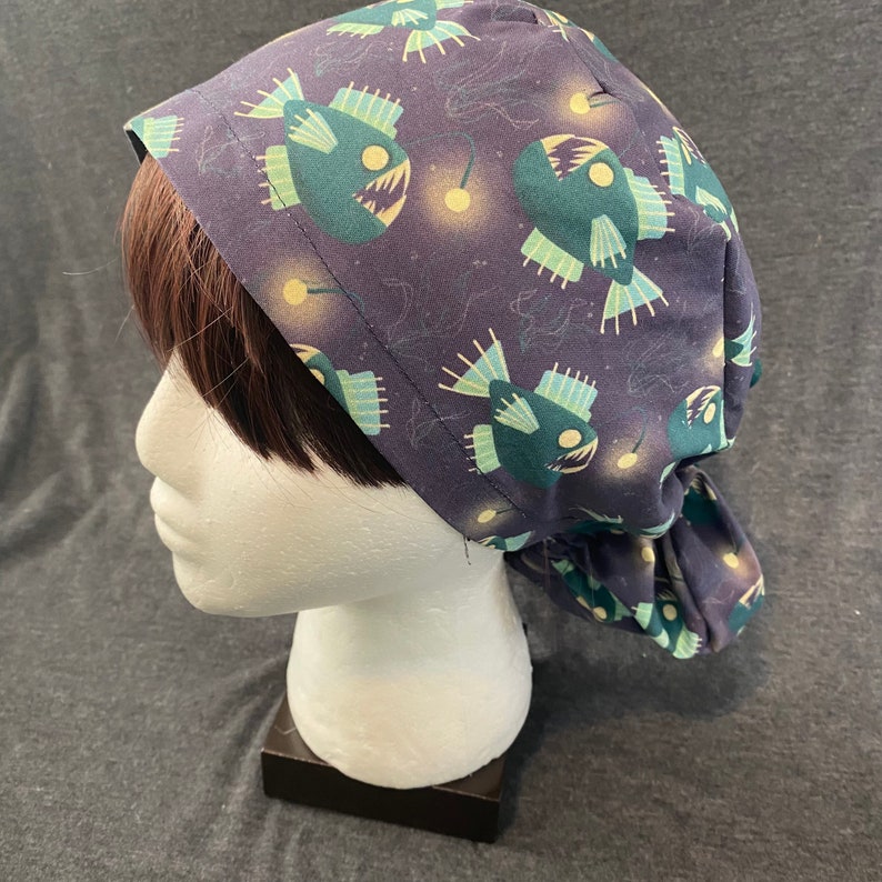 May include: A dark blue surgical scrub hat with a pattern of anglerfish and bioluminescent lights. The hat is tied at the back.