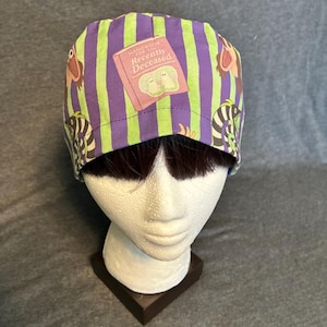 May include: A scrub cap featuring vertical purple and lime green stripes with a repeating pattern of a pink book titled Handbook for the Recently Deceased and cartoon characters. The fabric has a colourful print on a fitted, rounded head covering.