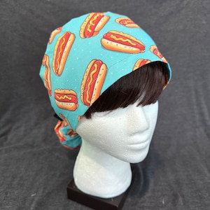 May include: A turquoise blue surgical scrub hat with a pattern of hot dogs. The hot dogs are yellow with red relish and mustard.