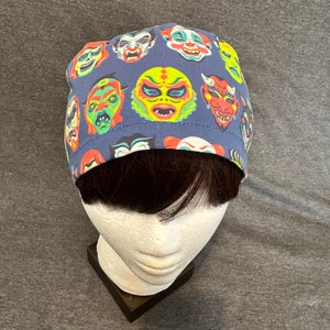 Puede incluir: Un gorro quirúrgico azul oscuro con un patrón repetido de caras de monstruos coloridos. Las caras son en tonos de rojo, verde, amarillo y blanco, creando un diseño lúdico. El gorro está hecho de una tela suave.
