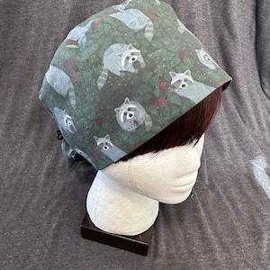 Puede incluir: Un gorro de quirófano verde con un patrón repetido de mapaches y bayas rojas. El gorro está hecho de una tela suave y cómoda y tiene un lazo en la parte posterior.