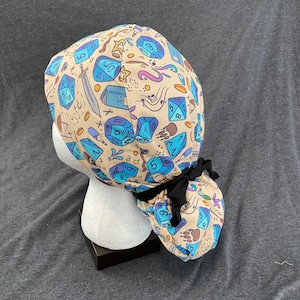 May include: A beige scrub hat with a black tie and a colorful pattern of dice and other symbols. The hat is on a white mannequin head.