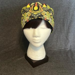 May include: A patterned scrub cap with a floral design in yellow, green, and orange on a black background. The cap is displayed on a white mannequin head with dark hair.