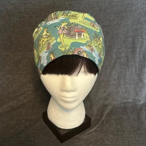 May include: A teal scrub cap featuring a whimsical frog-themed print. The fabric displays a repeating pattern of anthropomorphic frogs in a landscape with mushrooms and a small wooden structure. The cap is displayed on a mannequin head.