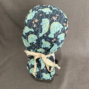 May include: A navy blue scrub cap featuring a repeating pattern of light blue manatees with yellow star accents, along with pink starfish and sprigs. The cap is tied at the back with a cream-colored ribbon.