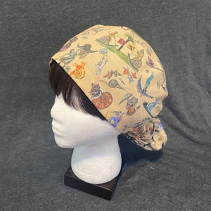 Peut inclure: Une toque de chirurgien beige avec un motif fantaisiste d'escargots, de personnages et d'illustrations. La toque est en tissu et est conçue pour être portée sur la tête. La toque est présentée sur une tête de mannequin blanche.