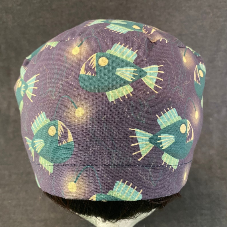 May include: A dark purple fabric scrub hat with a repeating pattern of anglerfish with glowing lures. The anglerfish are green and yellow.