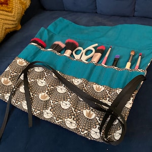May include: Teal fabric makeup roll with black ribbon ties and a brown and white patterned fabric with a repeated design of a crown and glasses. The roll is filled with makeup brushes and a pair of scissors.